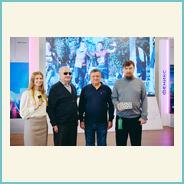 Сотрудники компании «ЭлекЖест» и участники фестиваля стоят на фоне сцены выставочного зала. Один из сотрудников держит в руках тифлофлешплеер. За ними на экране отображается изображение с массового спортивного мероприятия.